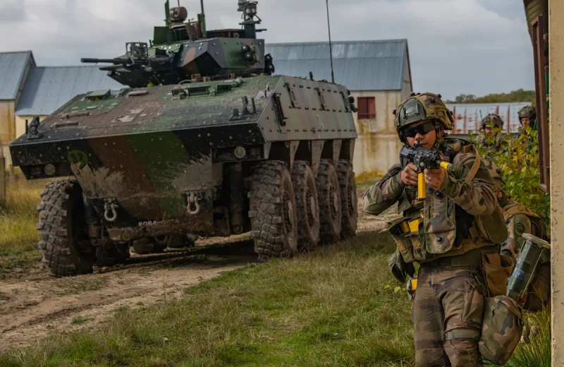 Des militaires en tenue camouflée à l’angle d’un mur, devant un véhicule blindé à roues.