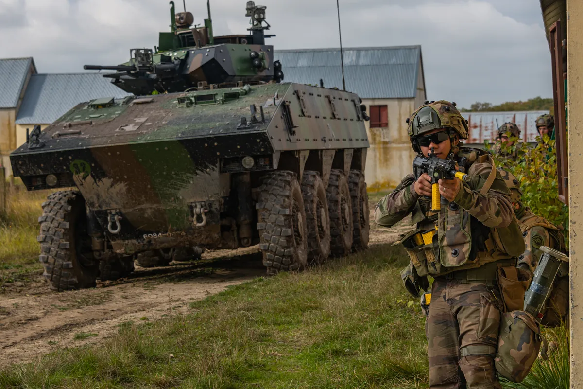 Des militaires en tenue camouflée à l’angle d’un mur, devant un véhicule blindé à roues.