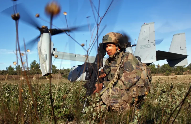 Un légionnaire accroupi sécurise une zone de poser, un MV-22B Osprey à l’arrière-plan.