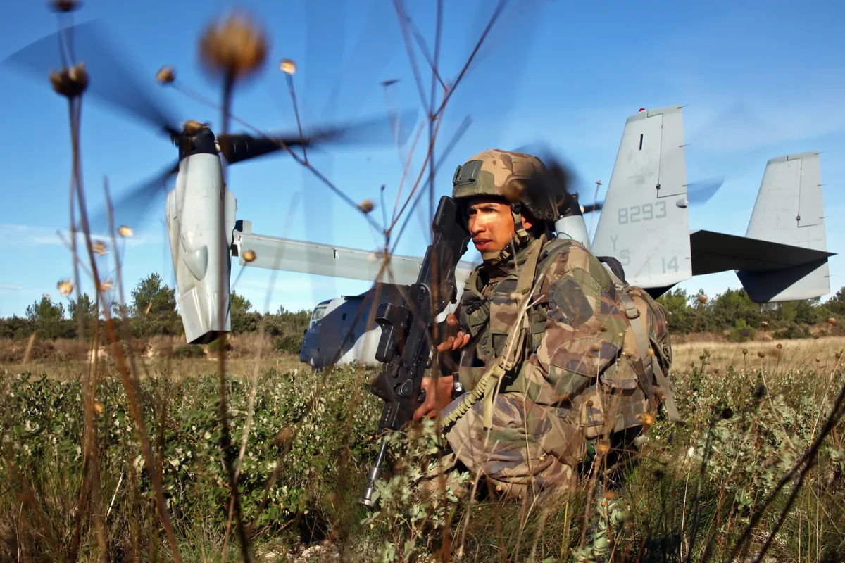 Un légionnaire accroupi sécurise une zone de poser, un MV-22B Osprey à l’arrière-plan.