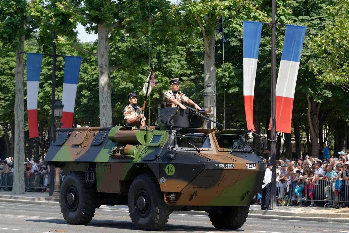 Un véhicule blindé à roues avec deux militaires sur le toit défile devant des spectateurs et des bannières tricolores.